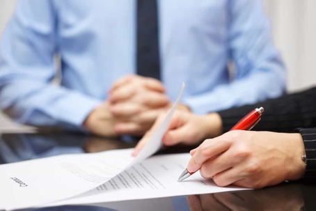 Close up of a person's hands signing a document with a businessman sitting patiently with hands crossed resting on the desk in the background